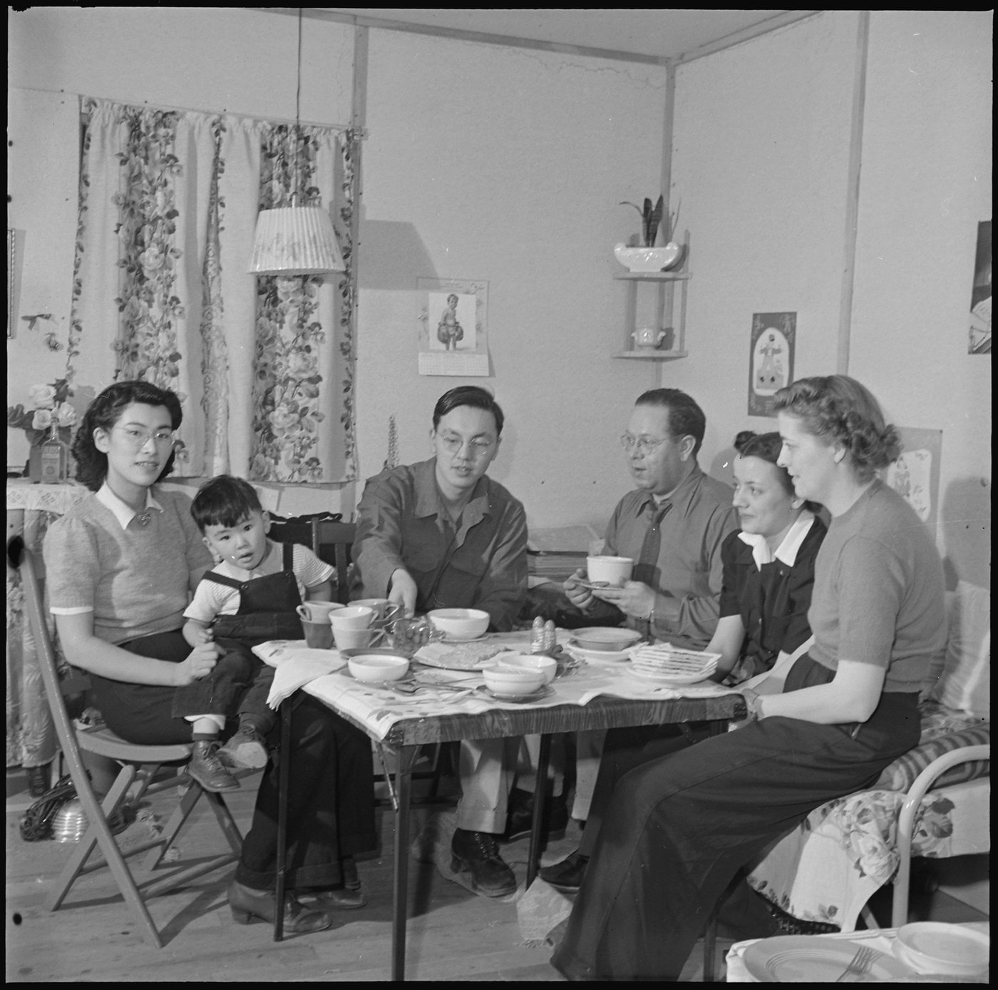 Scope and content:  The full caption for this photograph reads: Heart Mountain Relocation Center, Heart Mountain, Wyoming. In his barracks home at Block 7 - 21, Bill Hosokawa and his wife Alice serves oyster stew in an evening's visit with the members of the War Relocation Authority appointed personnel. Left to right is Julona Steinheider, High School Mathematics Teacher; Margaret Jensen, Center Librarian; Vaughn Mechau, Reports Officer; Bill, his son Mike and his wife, Alice. (This appears to have been written from a flipped image, but there's plenty of signs - the hands and watches being appropriate for right-handed people, and the calendar in the background, while not legible, looking more appropriate as it is now than in a mirror flip, that indicates this arrangement is correct).